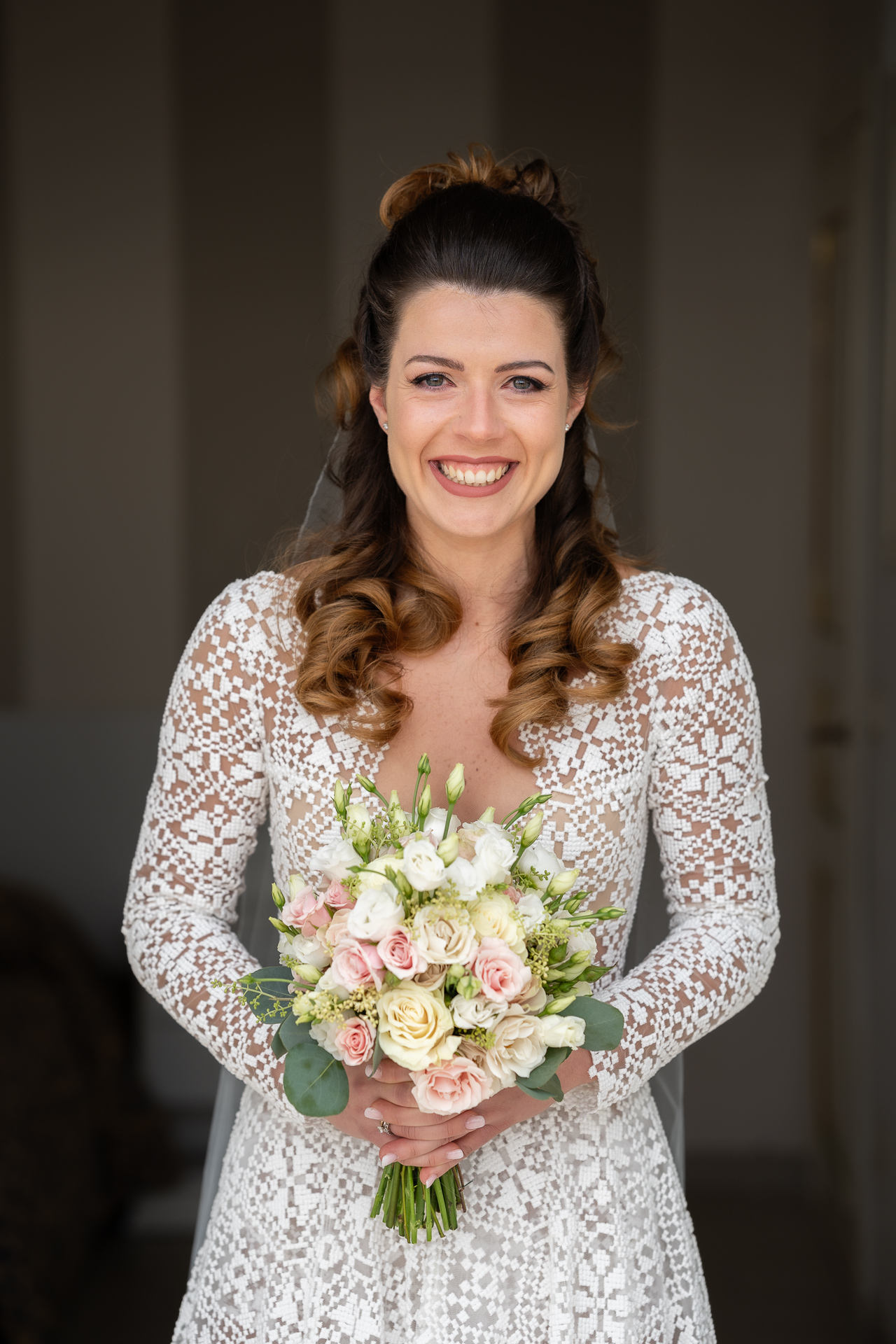 Una sposa con un'acconciatura raccolta sorride gioiosamente nella sua foto di matrimonio, tenendo in mano un mazzo di rose cipria e verde, indossando un abito di pizzo a maniche lunghe.