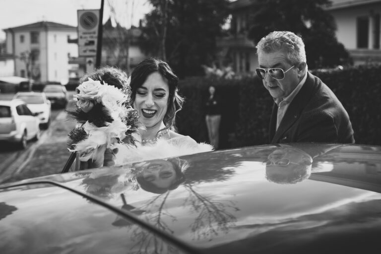 Una sposa gioiosa con un mazzo di fiori e un uomo anziano, forse suo padre, che escono da un'auto, con il loro riflesso visibile sulla superficie lucida dell'auto catturato in una bellissima fotografia.