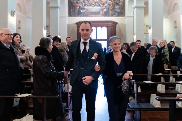 Uno sposo percorre la navata di una chiesa con al braccio una donna anziana, entrambi sorridenti, circondato da invitati elegantemente vestiti che li osservano in una cornice da foto matrimonio.