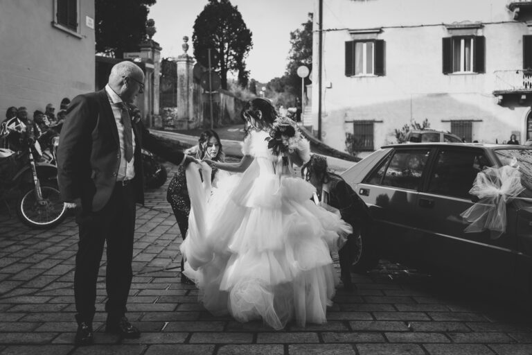 Una sposa in un voluminoso abito bianco con in mano un bouquet incontra un uomo distinto accanto a un'auto decorata, mentre un bambino guarda, in una scena di strada acciottolata durante il loro foto matrimonio.