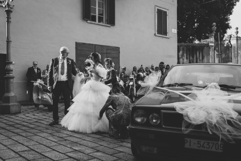 Una sposa con un voluminoso abito bianco riceve aiuto con il suo abito da due donne per strada, mentre gli ospiti guardano e un'auto adornata attende nelle vicinanze per il foto matrimonio.