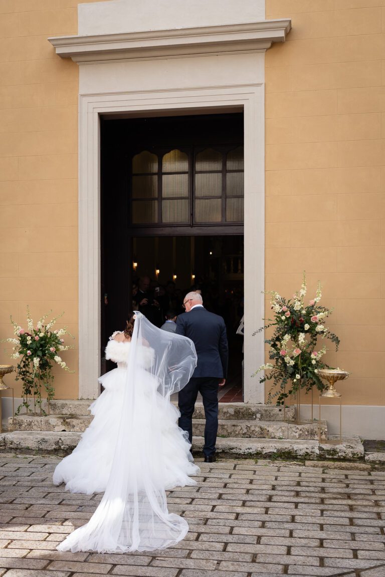 Una sposa con un fluido abito bianco e un velo viene scortata da un uomo anziano in abito formale in un luogo con una porta aperta, dove gli ospiti sono visibili all'interno. Accanto all'ingresso sono presenti delle composizioni floreali