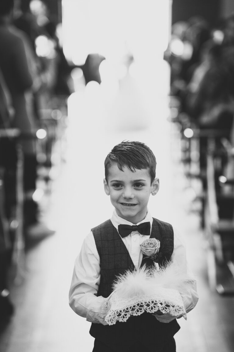 Un ragazzo in abiti formali, sorridente e con in mano un cuscino decorativo, si trova in una navata nuziale con gli ospiti sfocati sullo sfondo. Fotografia in bianco e nero.