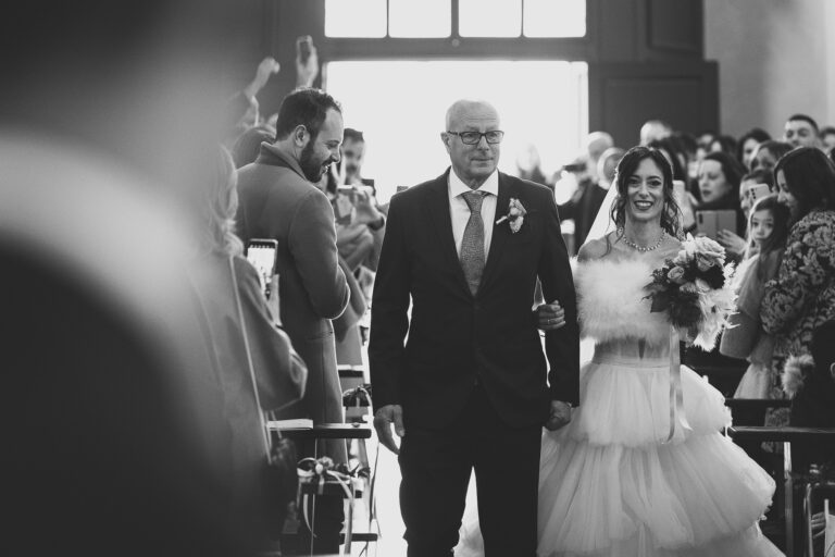 Una sposa in abito bianco sorride ampiamente mentre cammina lungo la navata con un uomo anziano in abito, entrambi circondati da ospiti in un luogo ben illuminato, catturando il momento perfetto per la fotografia