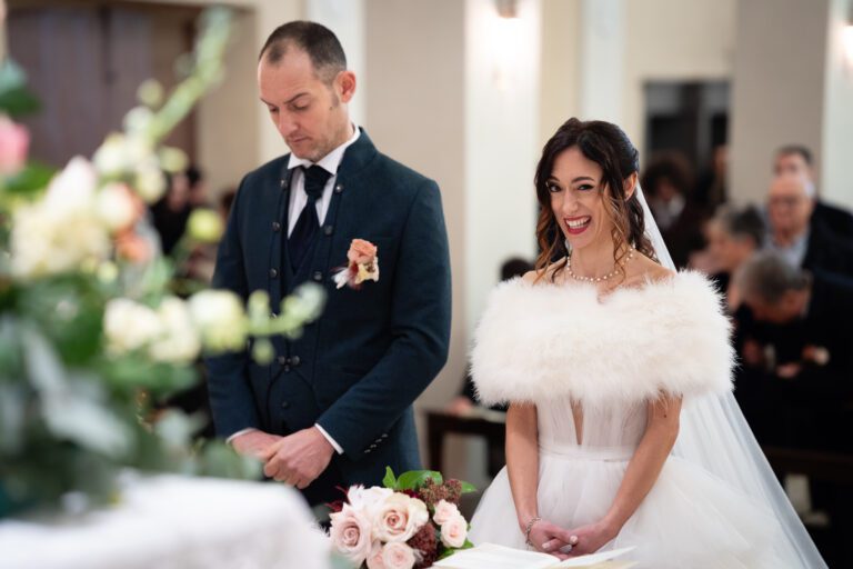 Una sposa in abito bianco e scialle di pelliccia sorride gioiosamente durante una cerimonia di matrimonio, mentre lo sposo accanto a lei, in abito scuro, guarda solennemente in basso. Sono all'altare
