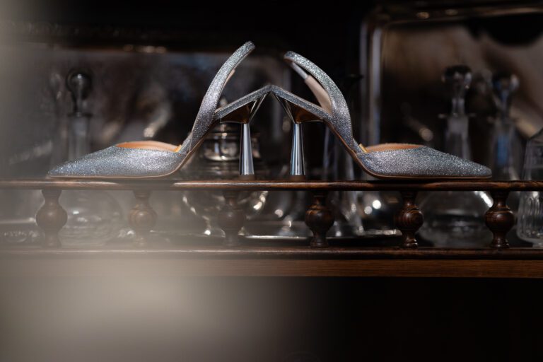 Un paio di scarpe col tacco argento scintillanti esposte su uno scaffale di legno, con uno sfondo scuro sfocato che esalta l'effetto glitter sulle calzature per un matrimonio fotografico.