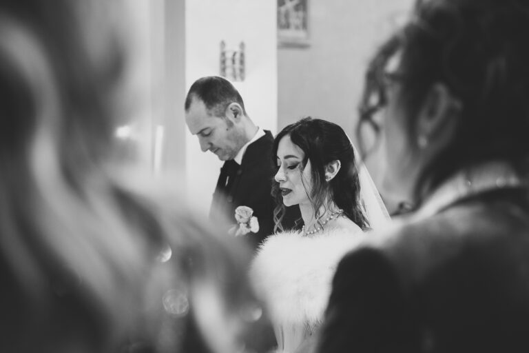 Una fotografía in bianco e nero di una sposa e uno sposo durante una cerimonia nuziale, visti tra le spalle degli invitati. Lo sposo guarda in basso mentre la sposa guarda avanti, entrambi vestiti formalmente.