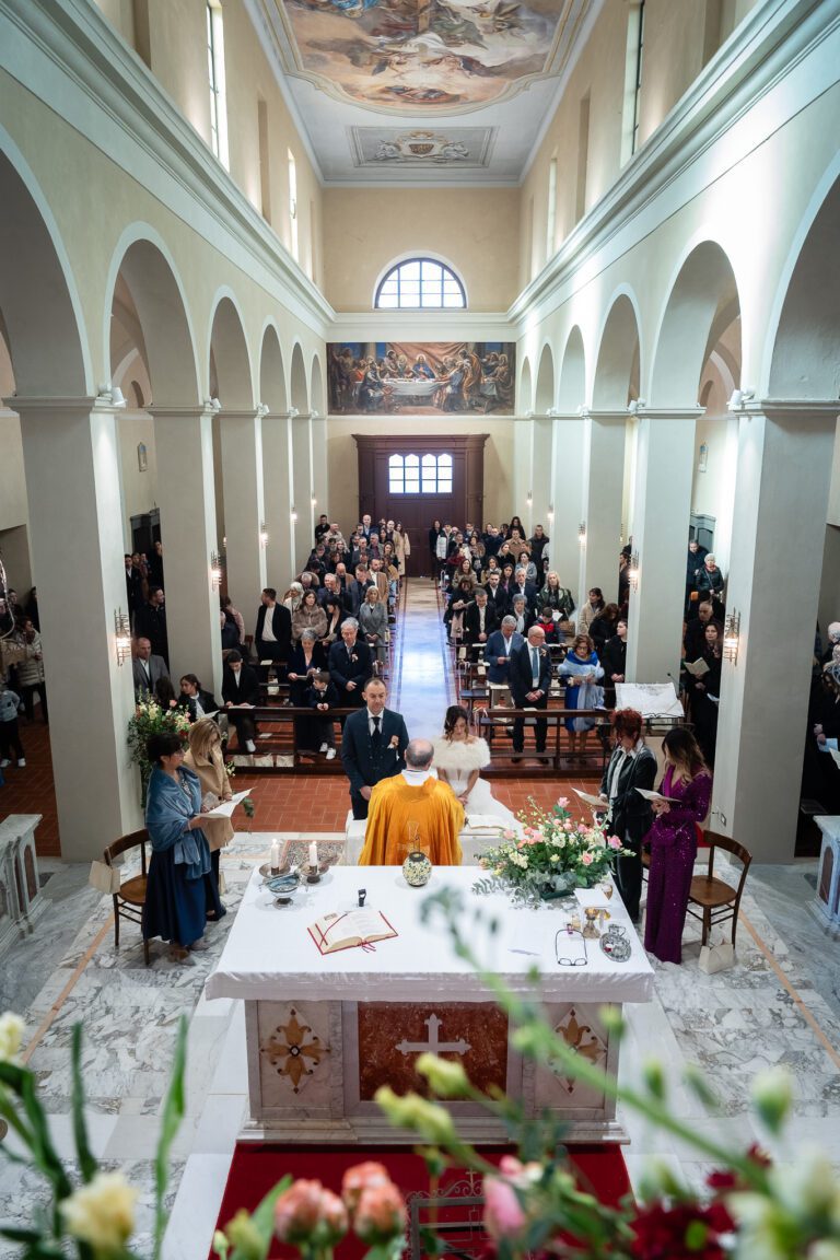 Veduta da dietro un altare, lungo la navata di una chiesa piena di ospiti che osservano gli sposi in piedi durante la cerimonia nuziale, catturati in una splendida fotografia.