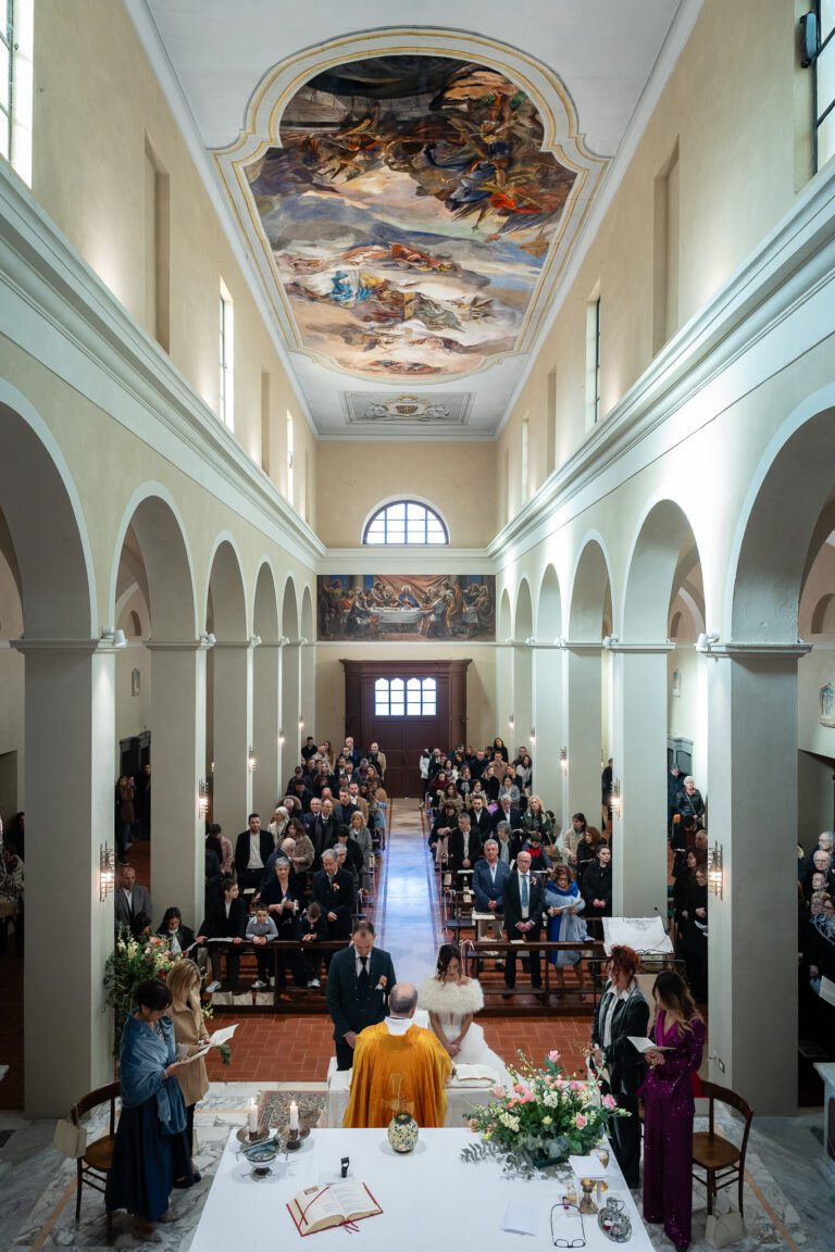 Una cerimonia di matrimonio all'interno di una grande sala dall'architettura elegante, porte ad arco e un vivace affresco sul soffitto. Gli ospiti sono seduti lungo i tavoli apparecchiati per il pasto, di fronte alla coppia in prima fila,