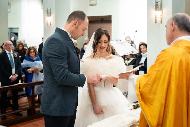 Gli sposi si scambiano gli anelli durante una cerimonia nuziale, catturati in una bellissima fotografia, circondati da ospiti, mentre un officiante in vesti gialle osserva. La sposa sorride con gioia, tenendo in mano un bianco peloso