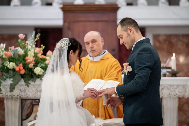 Gli sposi si scambiano gli anelli all'altare, sotto lo sguardo di un prete in vesti dorate, circondato da candele accese e decorazioni floreali, catturato in una bellissima fotografia.