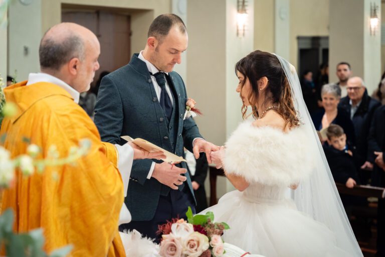 Gli sposi si scambiano gli anelli durante una cerimonia di matrimonio in una chiesa, con un prete e gli ospiti che osservano. La scena è catturata in una bellissima fotografia. La sposa indossa un abito bianco con stola di pelliccia