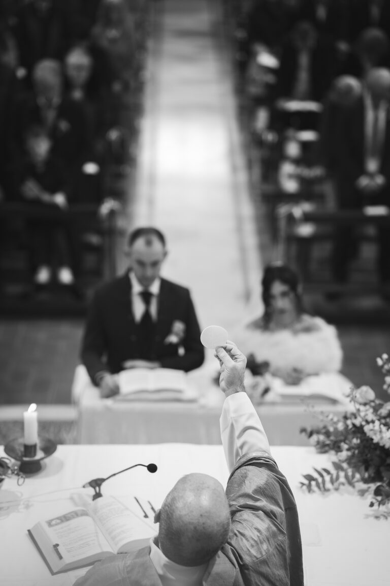 Un prete che tiene in mano un'ostia durante una cerimonia di matrimonio in una chiesa, con gli sposi seduti sullo sfondo e gli ospiti in piedi per la sessione fotografica.