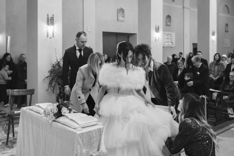 Una sposa alla cerimonia nuziale, circondata da ospiti e familiari, si aggiusta il suo voluminoso abito bianco assistita da tre donne in una chiesa caldamente illuminata per una fotografia.