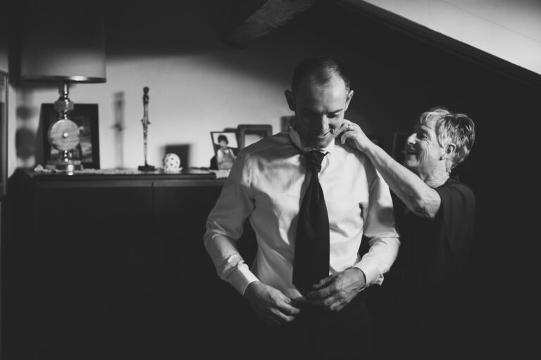 Una donna anziana aggiusta la cravatta di un uomo più giovane in una stanza caldamente illuminata con fotografie visibili e oggetti di un matrimonio sullo sfondo, trasmettendo un momento di cura e familiarità.