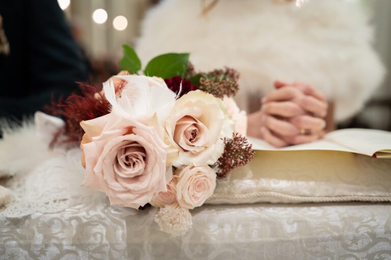 Una fotografia ravvicinata di un delicato bouquet da sposa con rose rosa e accenti piumati su un tavolo, con le mani della sposa giunte sullo sfondo.