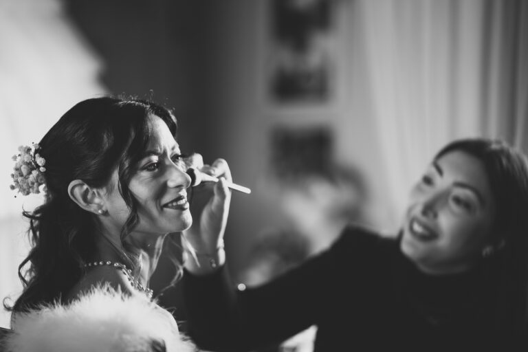 Una foto di matrimonio in bianco e nero di una donna che sorride mentre un'altra donna la trucca in una stanza caldamente illuminata. La donna truccata è adornata con un accessorio floreale per capelli.