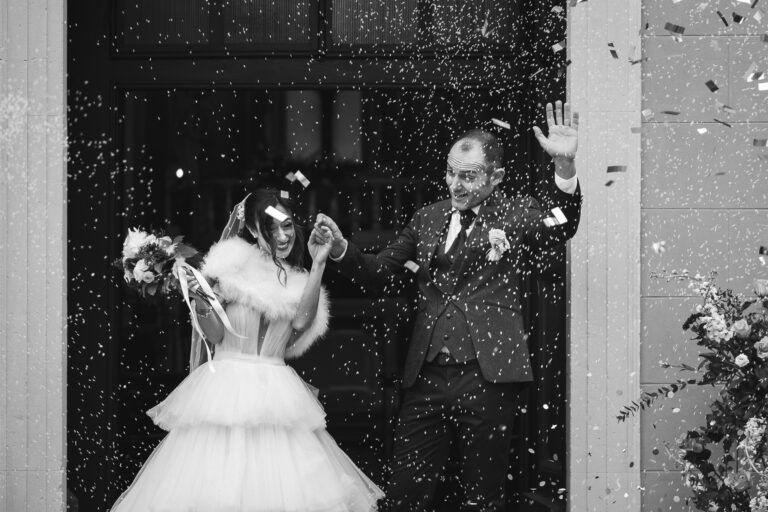 Una fotografia in bianco e nero di una sposa e uno sposo gioiosi che escono da un edificio mentre vengono inondati di coriandoli.