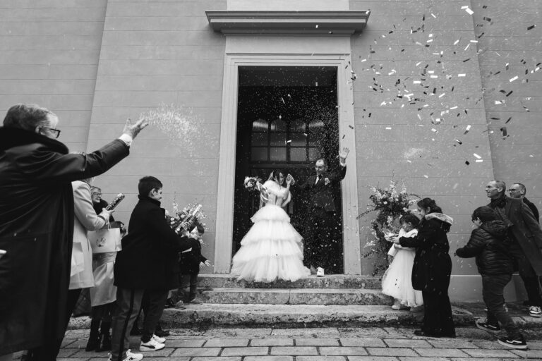 Una coppia di sposini esce da un edificio accolta dagli ospiti che lanciano coriandoli e riso. La sposa, in abito bianco, e lo sposo festeggiano in mezzo al caos gioioso catturato in una splendida fotografia.
