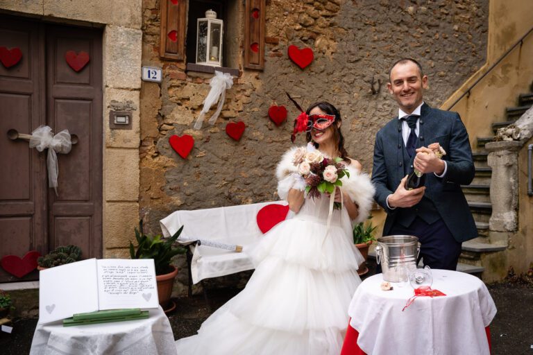 Una sposa gioiosa in abito bianco e maschera rossa tiene in mano un bouquet, in piedi accanto a uno sposo in abito, con in mano una maschera, vicino a un tavolo decorato con un libro degli ospiti, sullo sfondo di un vecchio