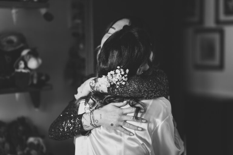 Due persone si stringono in un abbraccio sentito, una con un accessorio floreale tra i capelli, in una stanza addobbata per un matrimonio con oggetti vari, trasmettendo in bianco e nero un momento di affetto.