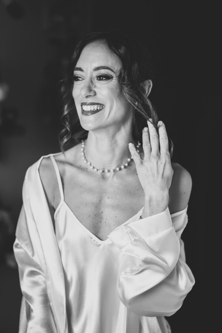 Una foto in bianco e nero di una donna sorridente in una veste di seta, che si tocca i capelli con una collana di perle al collo. Appare gioiosa ed elegante al suo matrimonio.