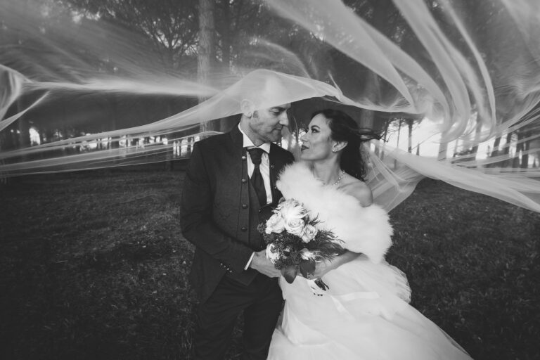 Una fotografia in bianco e nero di una sposa e uno sposo che si sorridono, avvolti in un velo fluente con alberi sfocati sullo sfondo.