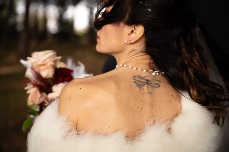 Una sposa con un delicato tatuaggio a farfalla sulla schiena, che indossa una collana di perle e una stola di pelliccia bianca, con in mano un bouquet, vista da un angolo laterale ravvicinato in questa bellissima fotografia.