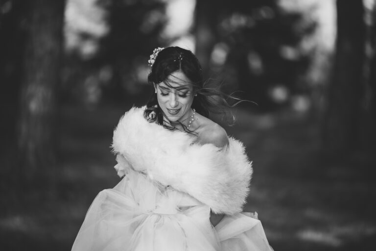 Una foto di matrimonio in bianco e nero di una sposa all'aperto, che si aggiusta il vestito e guarda verso il basso. Indossa una tiara, orecchini e una stola di pelliccia sulle spalle.