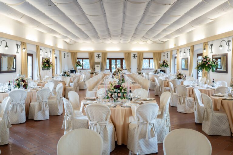 Elegante sala per ricevimenti di nozze con tavoli splendidamente decorati, coprisedie bianche e centrotavola floreali, evidenziati dalla luce naturale proveniente dalle grandi finestre catturate in una *foto matrimonio*.