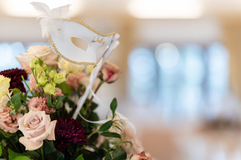 Un'elegante maschera bianca da ballo ornata con piume e finiture dorate, etichettata "dulce", è messa a fuoco sopra un mazzo di fiori assortiti in una foto di matrimonio, con uno sfondo leggermente sfocato