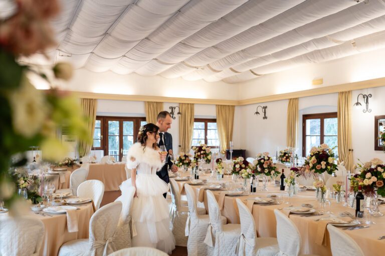 Una coppia di sposini entra in una sala da pranzo elegantemente arredata con centrotavola floreali e tavole apparecchiate, raggiante di gioia per il loro foto matrimonio.