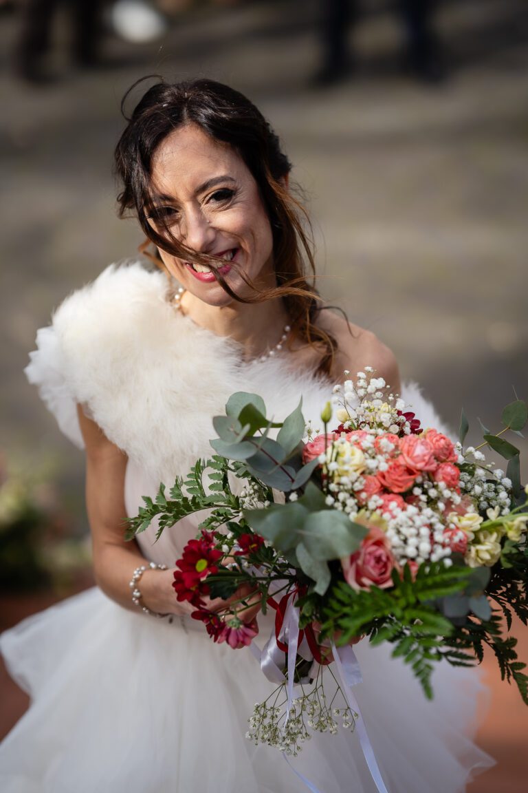Una sposa gioiosa in abito bianco e scialle di pelliccia, con in mano un mazzo di fiori rosa, rossi e bianchi, sorride brillantemente in una foto di matrimonio in una giornata soleggiata.