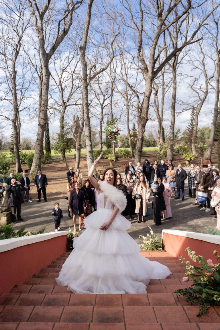 Una sposa lancia con gioia il suo bouquet verso una folla di invitati in un ambiente esterno fiancheggiato da alberi alti e spogli sotto un cielo limpido durante un foto matrimonio.