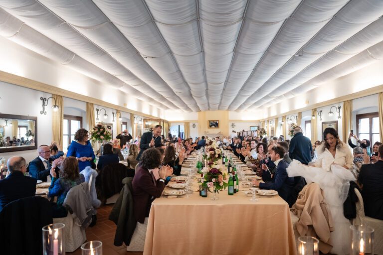 Un movimentato ricevimento di nozze con gli ospiti seduti su lunghi tavoli ornati da decorazioni floreali, sotto un soffitto drappeggiato di bianco. In un gioioso foto matrimonio, gli sposi interagiscono con i partecipanti.