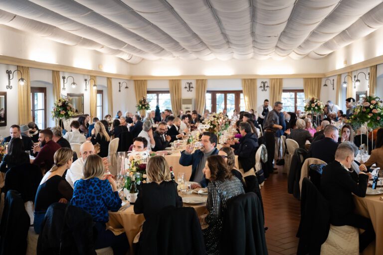 Una vivace sala banchetti con ospiti elegantemente vestiti seduti attorno a tavoli rotondi adornati con centrotavola floreali, impegnati in una conversazione, durante un evento di matrimonio formale.