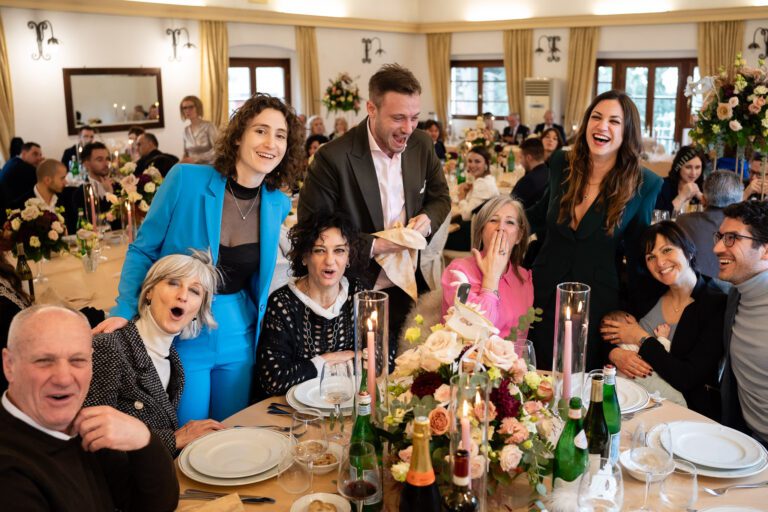 Un gruppo gioioso di persone ad una "foto matrimonio", ridendo e interagendo attorno ad un tavolo decorato con fiori. Un uomo in giacca e cravatta intrattiene la folla con un discorso.