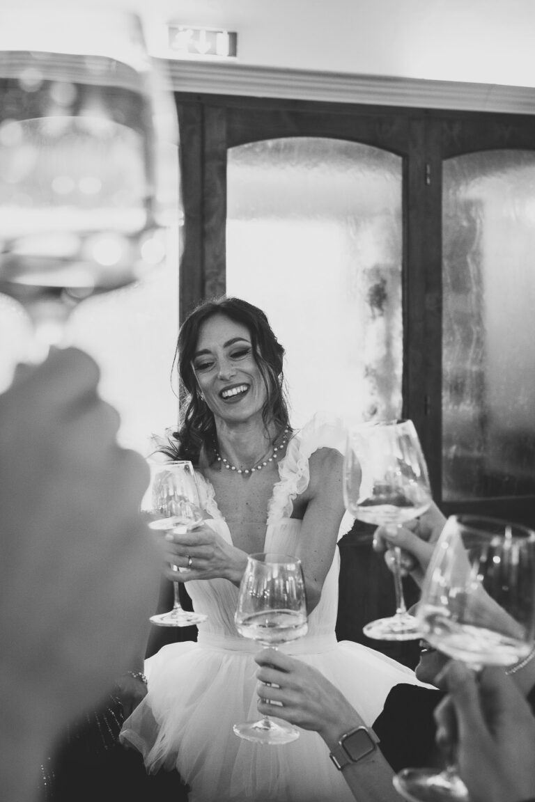 Una gioiosa sposa in abito bianco ride mentre brinda con gli ospiti, tenendo in mano un bicchiere di vino in una stanza poco illuminata e dall'arredamento elegante durante il suo foto matrimonio.