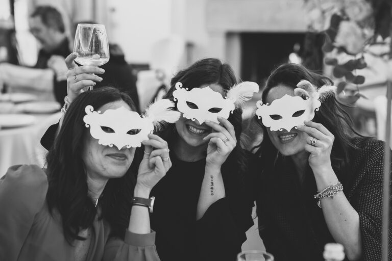 Tre donne durante un servizio fotografico di matrimonio, sorridono e tengono in mano maschere stravaganti, con un uomo sfocato che tiene un bicchiere di vino sullo sfondo.