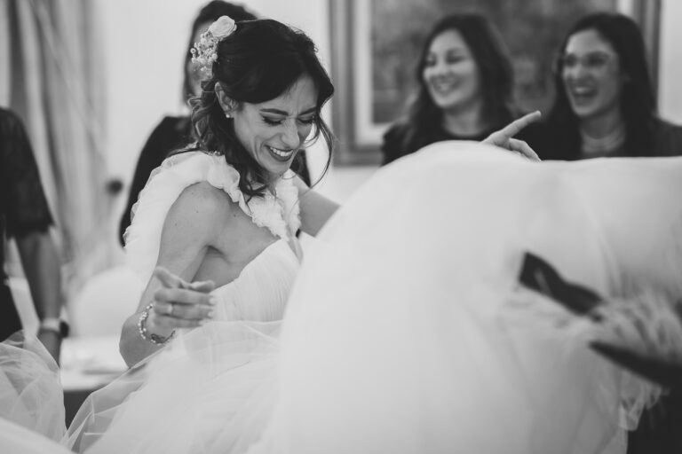 Una sposa gioiosa in abito bianco ride mentre volteggia, circondata da ospiti sorridenti in una stanza ben illuminata, catturando un momento di felicità durante il suo matrimonio.