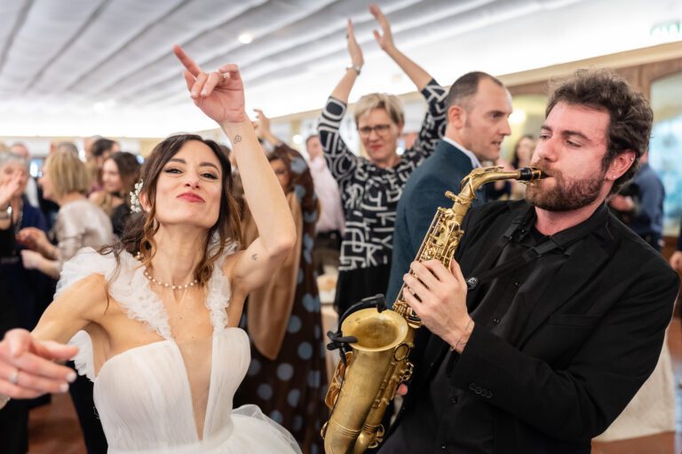 Una sposa balla con gioia, puntando il dito verso l'alto, accompagnata da un sassofonista che suona energicamente durante un vivace ricevimento di nozze.