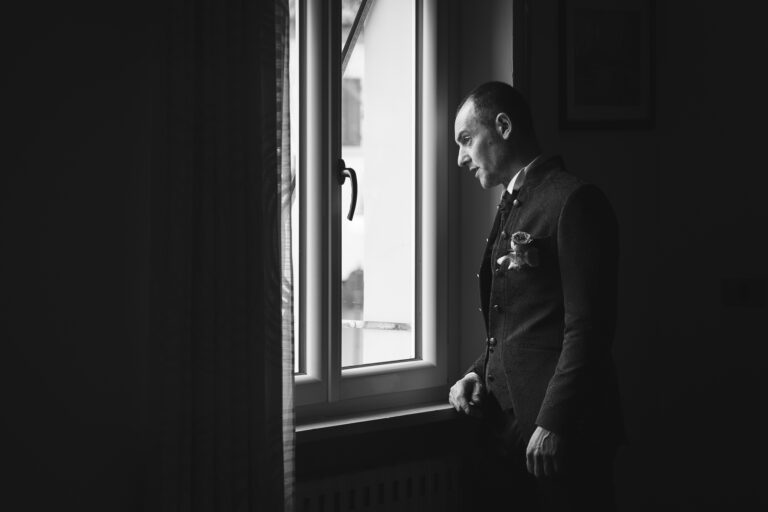 Un uomo con un gilet formale guarda pensieroso fuori da una finestra, evidenziando un momento di introspezione in una foto di matrimonio in bianco e nero.