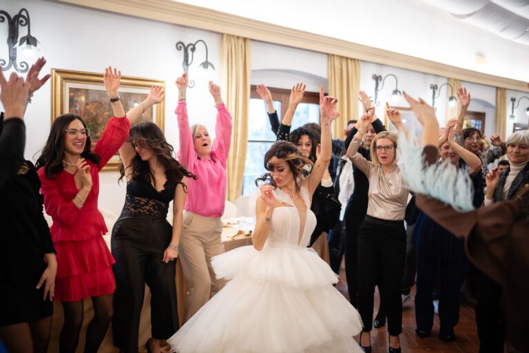 Una sposa in abito bianco balla gioiosamente circondata da un gruppo di invitati, tutti alzano le mani e applaudono in una stanza decorata con dipinti. Questa foto matrimonio cattura perfettamente la felicità e l'eccitazione.