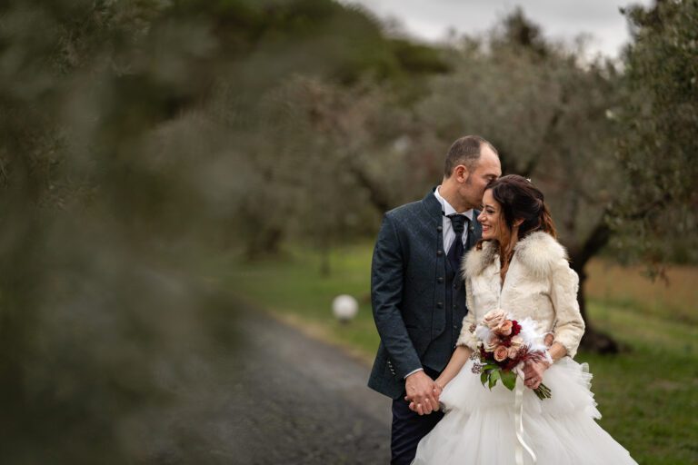 In una foto matrimonio, gli sposi si tengono per mano e sorridono, in piedi su un sentiero immerso nel verde, con lo sposo che sussurra all'orecchio della sposa. La sposa indossa un bianco