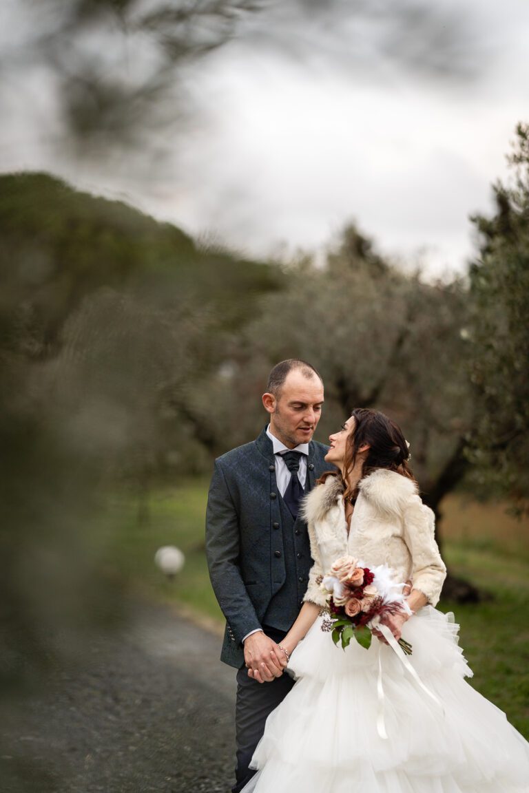 Una coppia di sposini si tiene per mano e si sorride in un ambiente sereno all'aperto, circondato da alberi, con la sposa che indossa un abito bianco e una stola di pelliccia e lo sposo in un abito blu. Questo