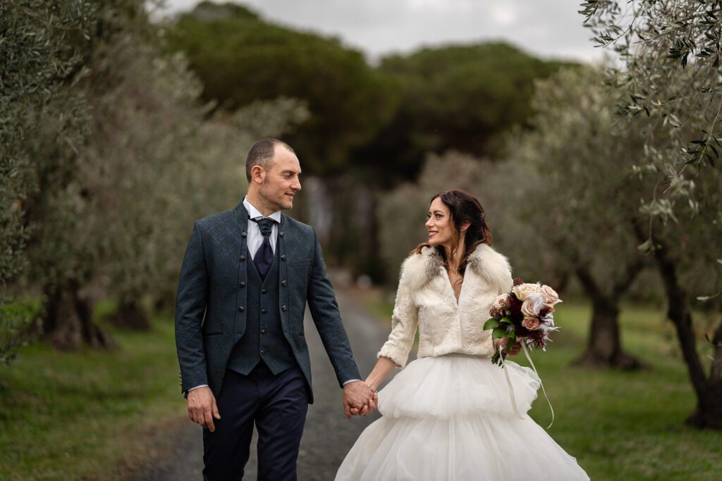 Una foto di matrimonio mostra una sposa ed uno sposo che si tengono per mano, camminando lungo un viale fiancheggiato da ulivi e guardandosi
