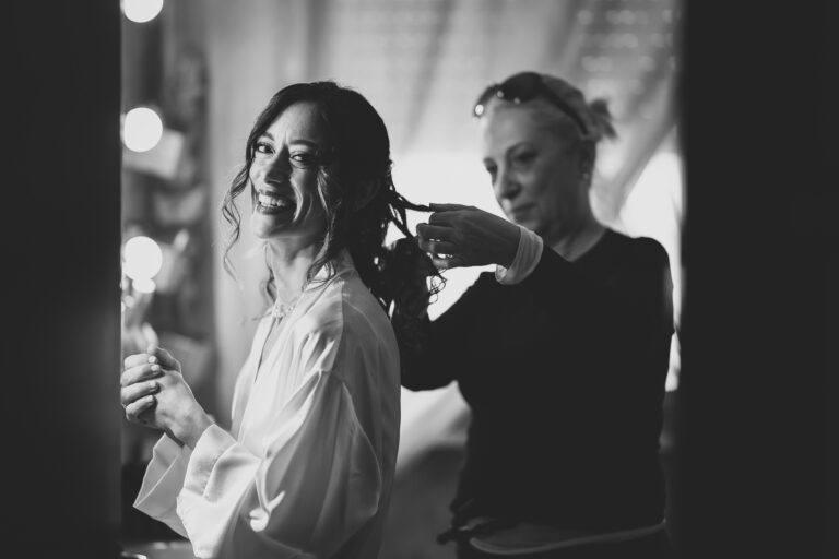 Una foto di matrimonio in bianco e nero di una donna sorridente in vestaglia che si fa acconciare i capelli da un'altra donna in un salone, con luci intense e specchi sullo sfondo.