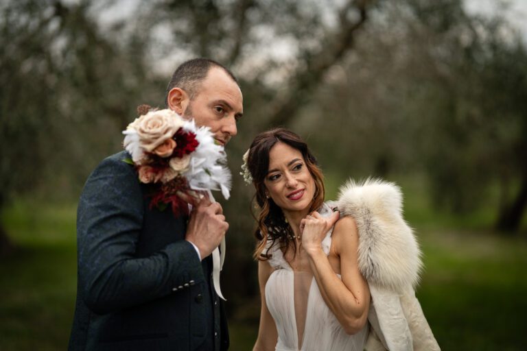 Una sposa e uno sposo in posa giocosamente all'aperto con lo sposo che sbircia da dietro un grande bouquet floreale. La sposa, che indossa un abito bianco e una stola di pelliccia, sorride alla telecamera in un boschetto di