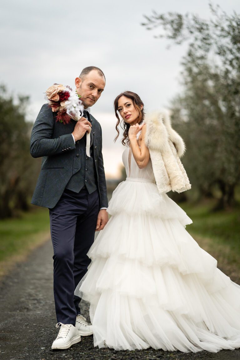 Una foto di matrimonio che mostra una sposa e uno sposo con un bouquet, camminando su un sentiero rurale tra gli ulivi; la sposa indossa un ab