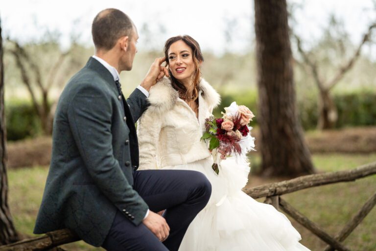 Una sposa in abito bianco e scialle di pelliccia siede su una staccionata di legno, con in mano un bouquet, mentre uno sposo in abito blu le tocca delicatamente il viso. Sono circondati da un sereno uliveto